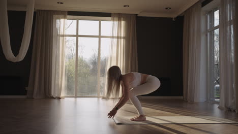 A-young-beautiful-woman-in-a-white-suit-practices-yoga-in-slow-motion-in-the-sun-in-a-hall-with-large-windows.-Sport-and-healthy-lifestyle